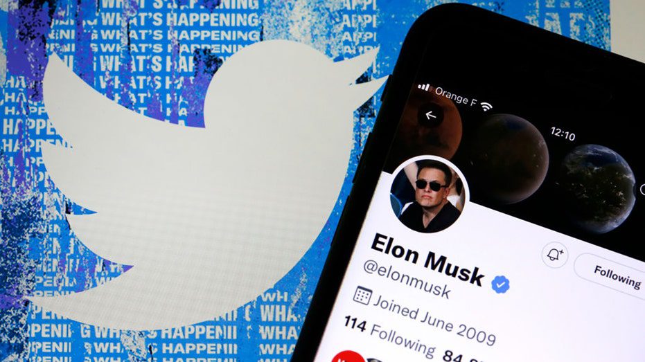 Elon Musk's Twitter account is displayed on the iPhone screen in front of the homepage of the Twitter website