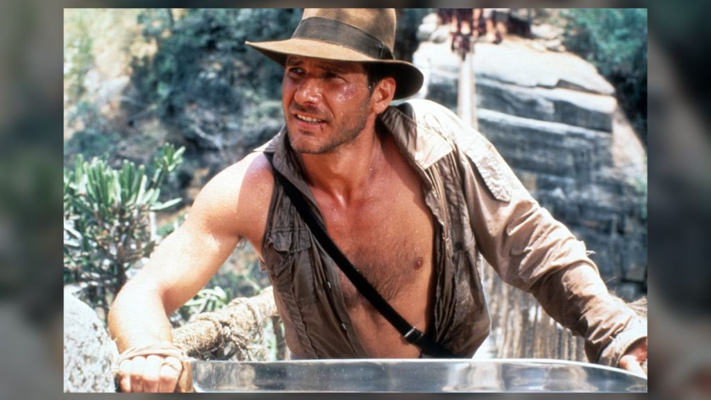 Harrison Ford wears his iconic Indiana Jones hat and an open jacket missing a sleeve.