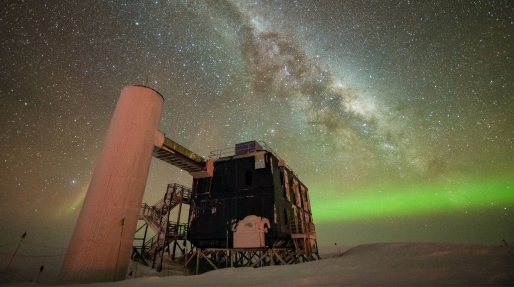 The IceCube Neutrino Observatory is seen under a starry night sky, with the Milky Way appearing over low auroras in the background.